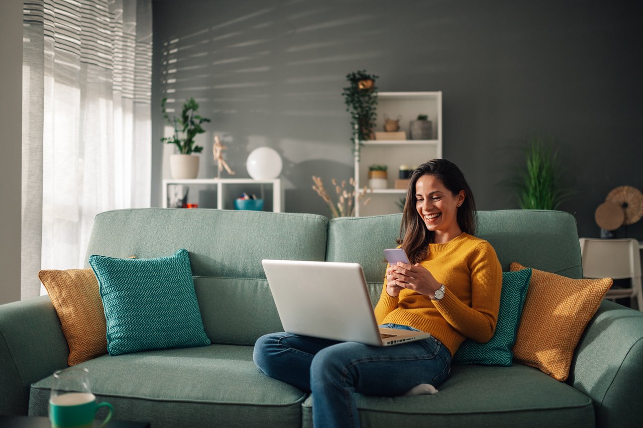 <p>Smiling caucasian woman using laptop and typing on a phone while sitting on couch at home. Happy female shopping or chatting online in social network, typing blog, sending message or email. Copy space</p>